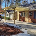 Stone pavers are being laid along a brick house, pergola under construction