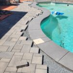 Backyard pool area under renovation with workers laying grey pavers along a curved edge near the pool.